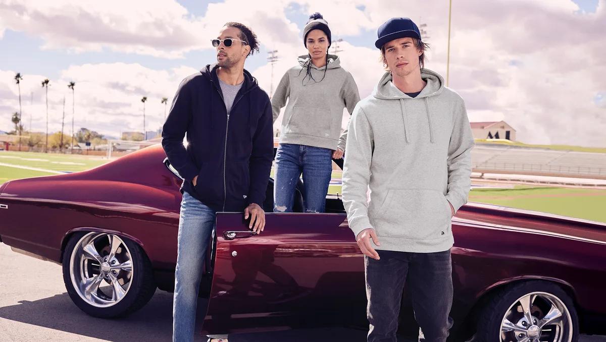 Three people in New Era fleece hoodies and beanies beside a classic car